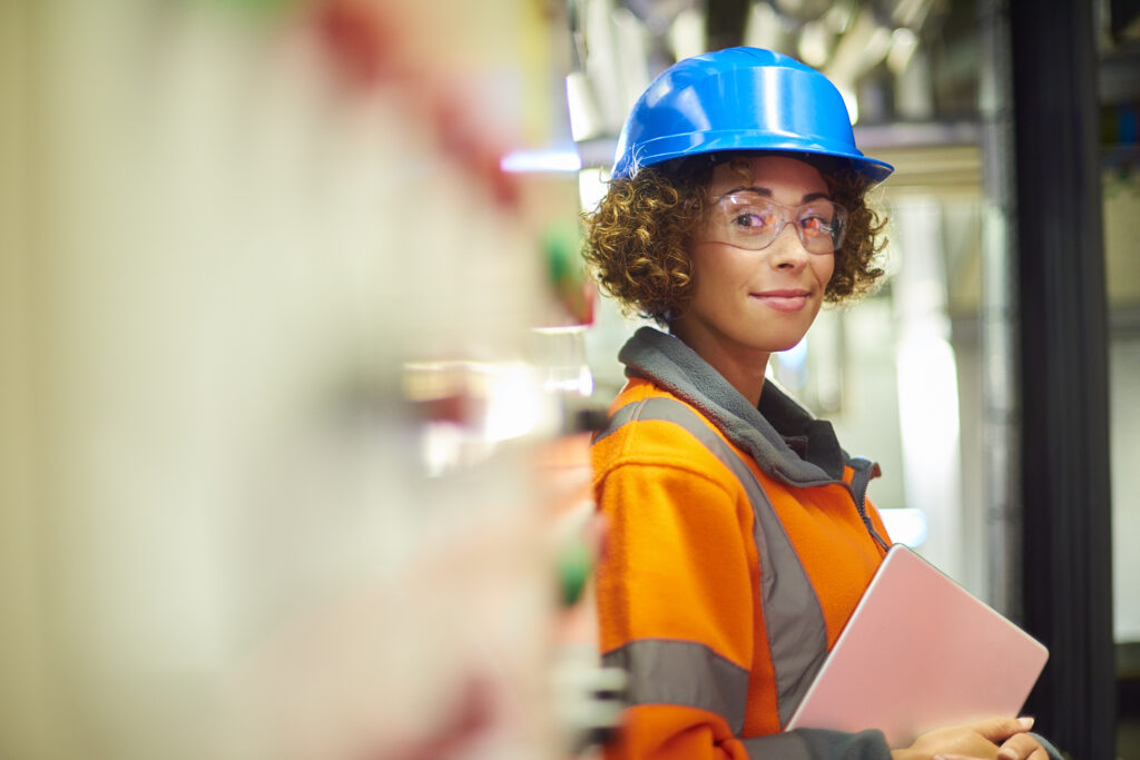 A female industrial service engineer has just conducted a safety check of a control panel in a boiler room. She is wearing hi vis, hard hat, safety glasses and holding a digital tablet that she used to conduct a safety inspection. The engineer is stated next to the control panel looking to camera and smiling.